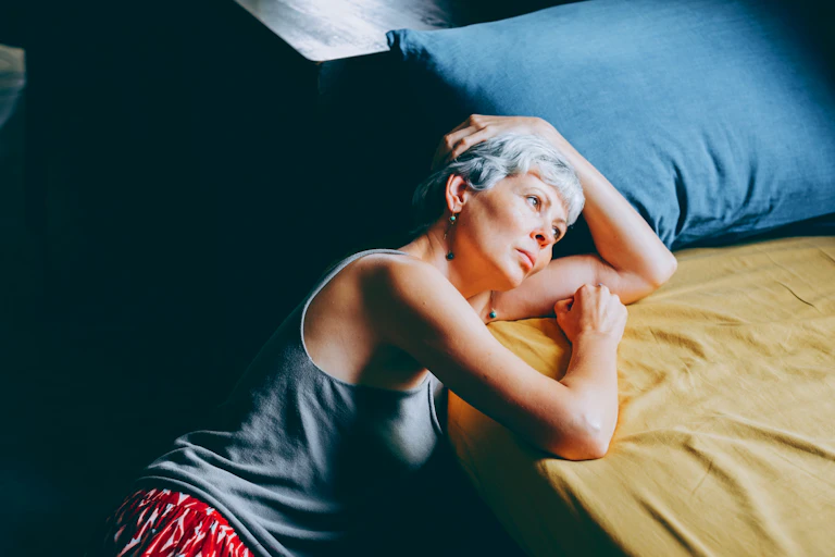 Woman with grey hair and dangling earrings leans against her bed