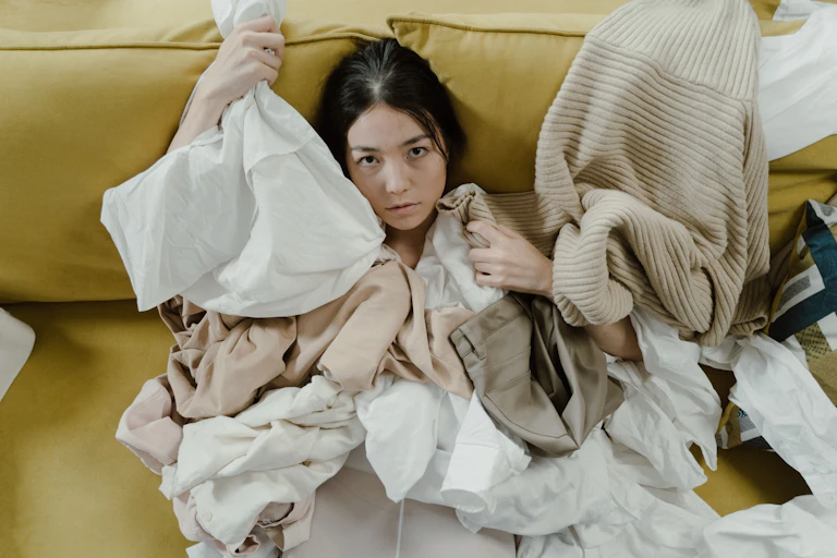 A young girl on a couch has clothing strewn around her.