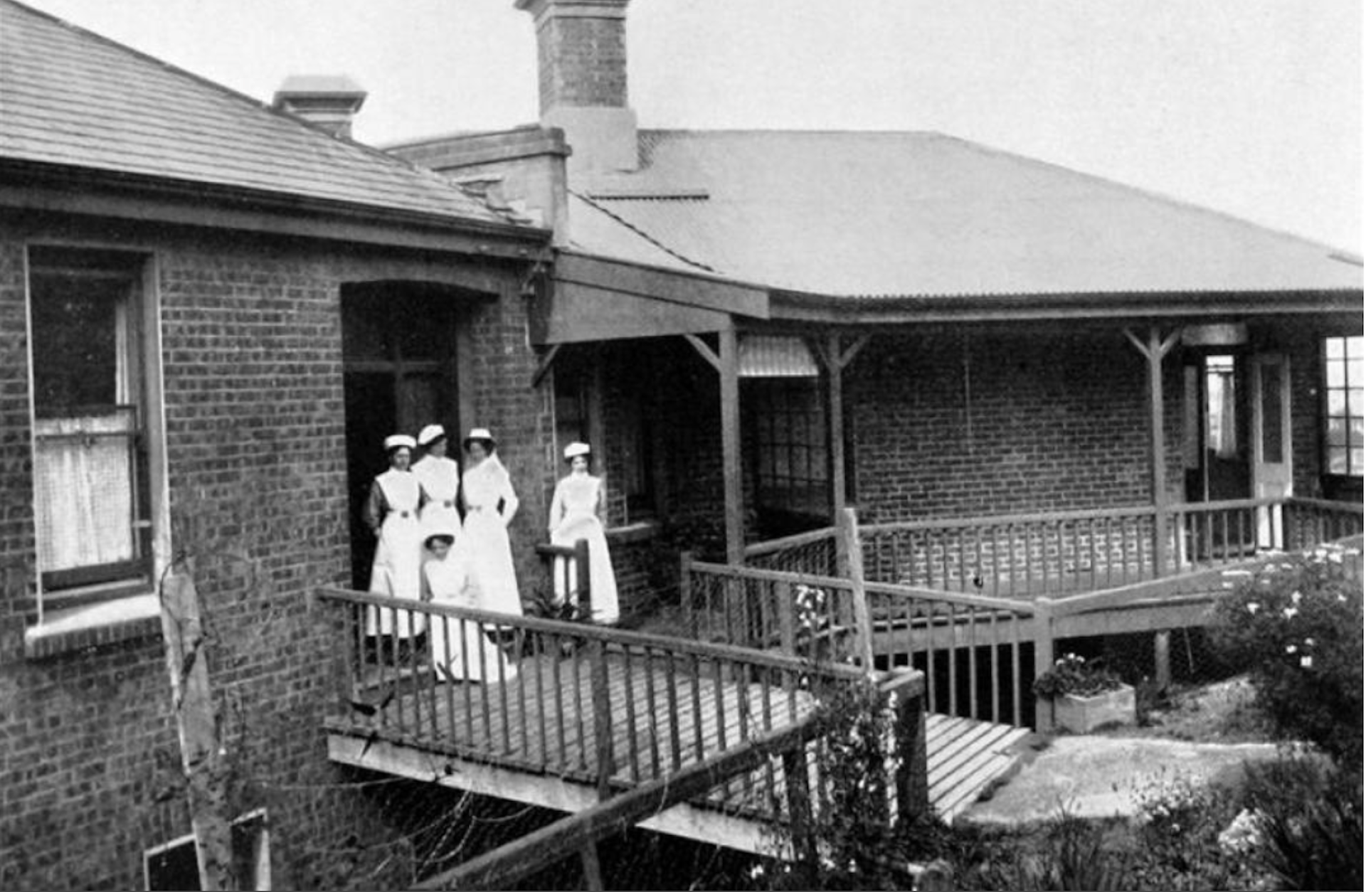 A historic black-and-white photograph shows a group of five nurses wearing vintage white uniforms and caps on an elevated wooden balcony outside a brick hospital building.