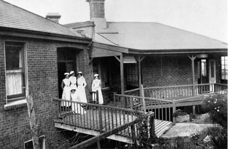 A historic black-and-white photograph shows a group of five nurses wearing vintage white uniforms and caps on an elevated wooden balcony outside a brick hospital building.