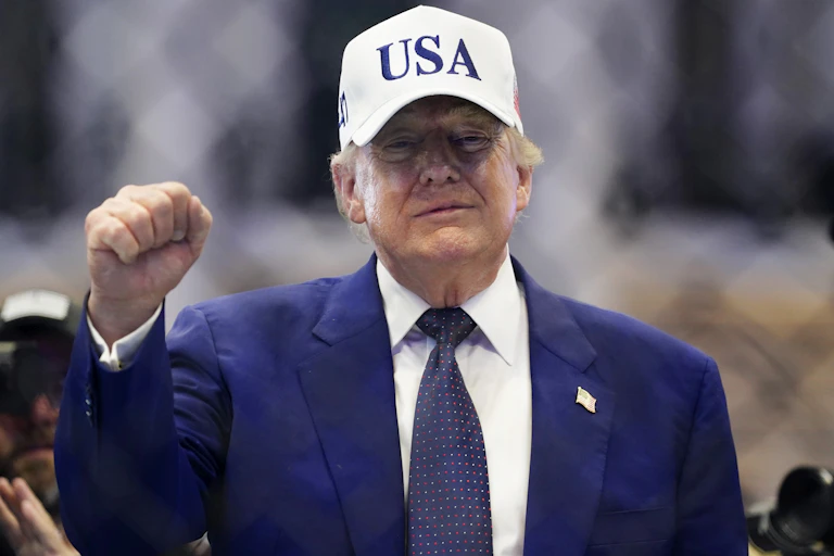 US president, Donald Trump, holds up his first while wearing a USA baseball cap.