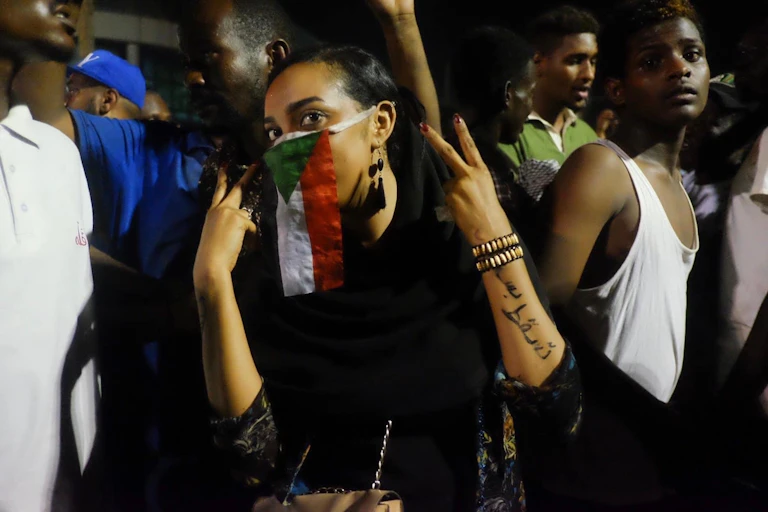 A woman holding up her hands in the peace sign while covering up half her face with a flag that has black, white, red and green stripes