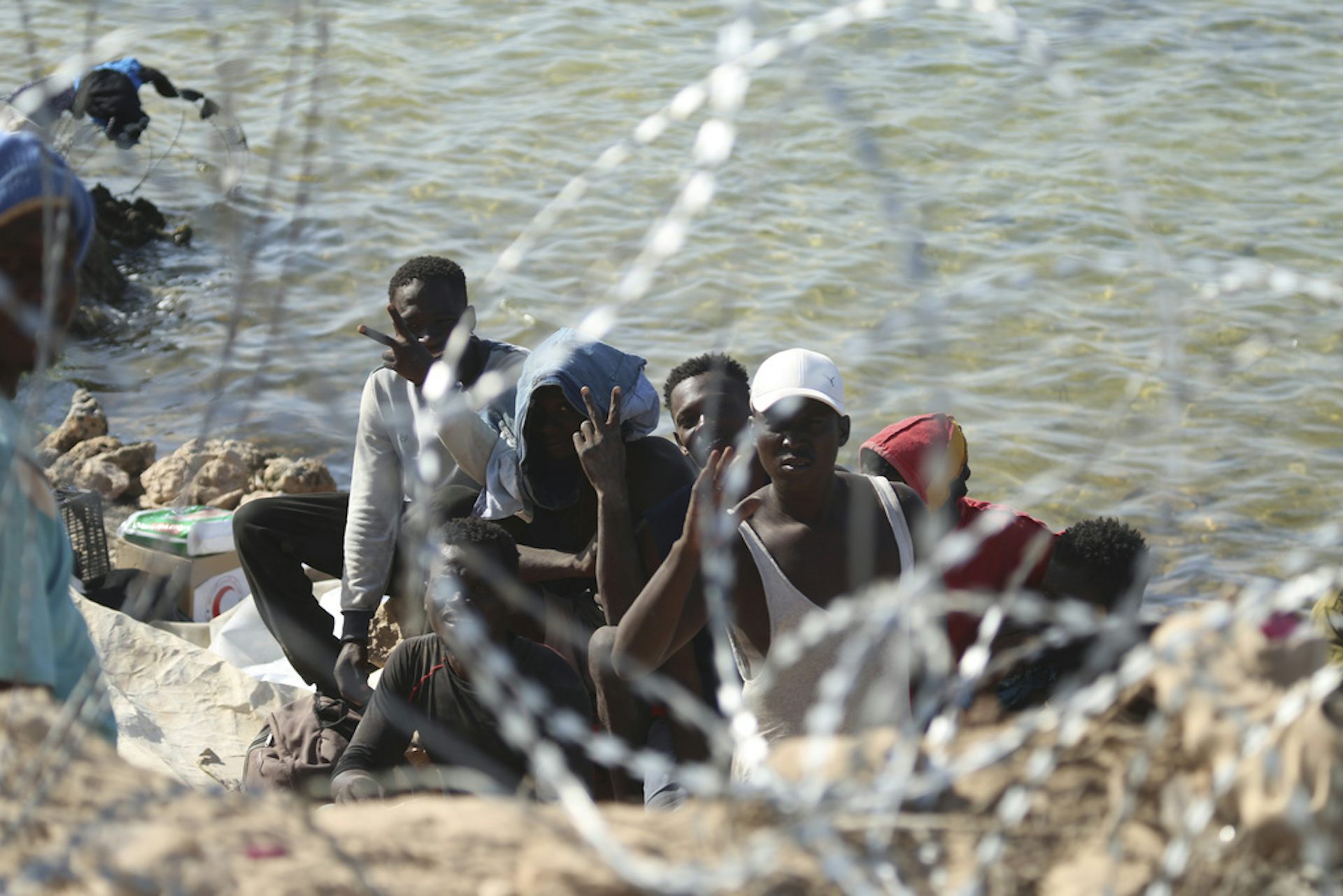 People gather by a shore with barbed wire in the foreground.
