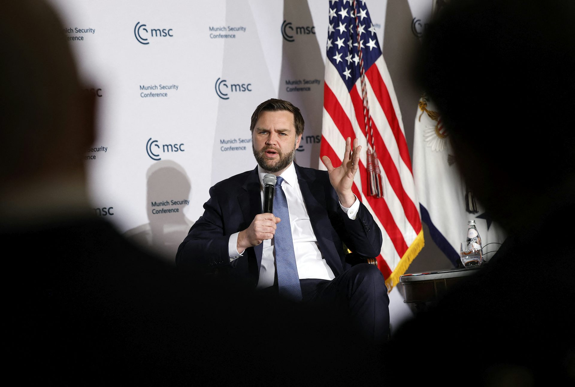 JD Vance assis sur scène tenant un micro devant un drapeau américain