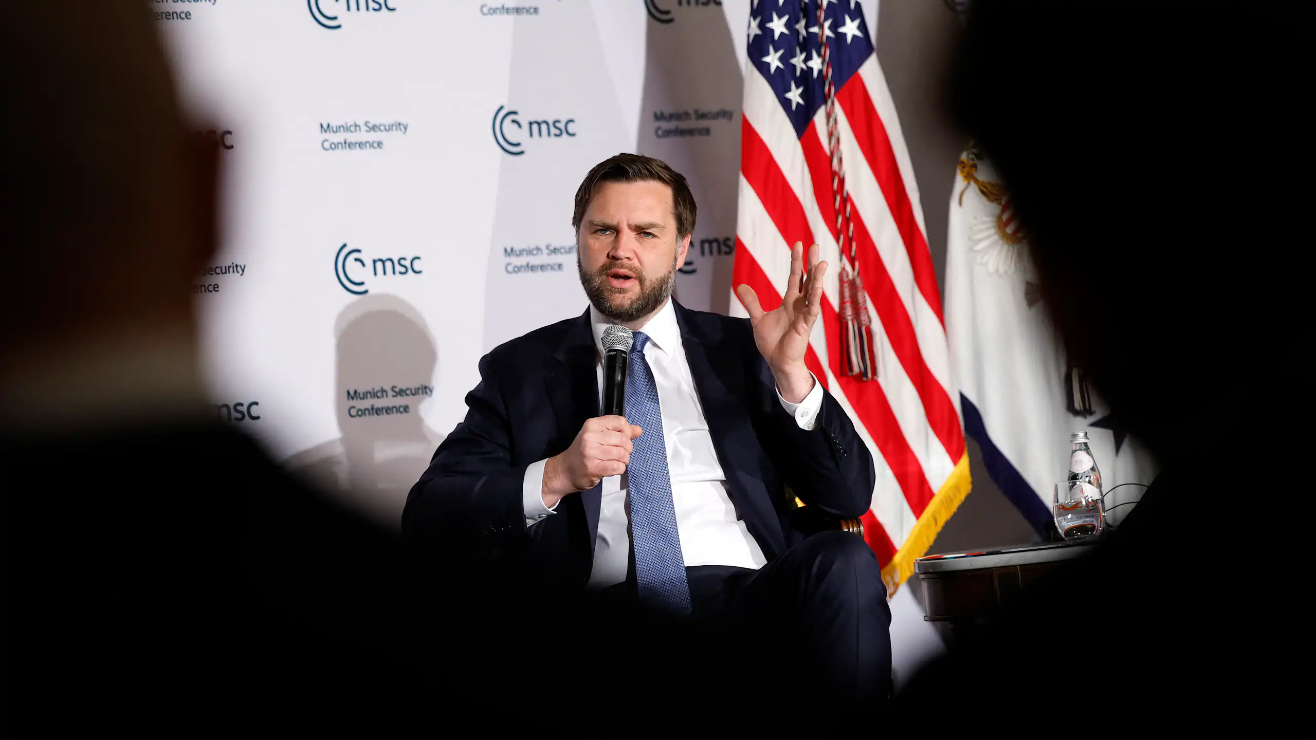 JD Vance assis sur scène tenant un micro devant un drapeau américain