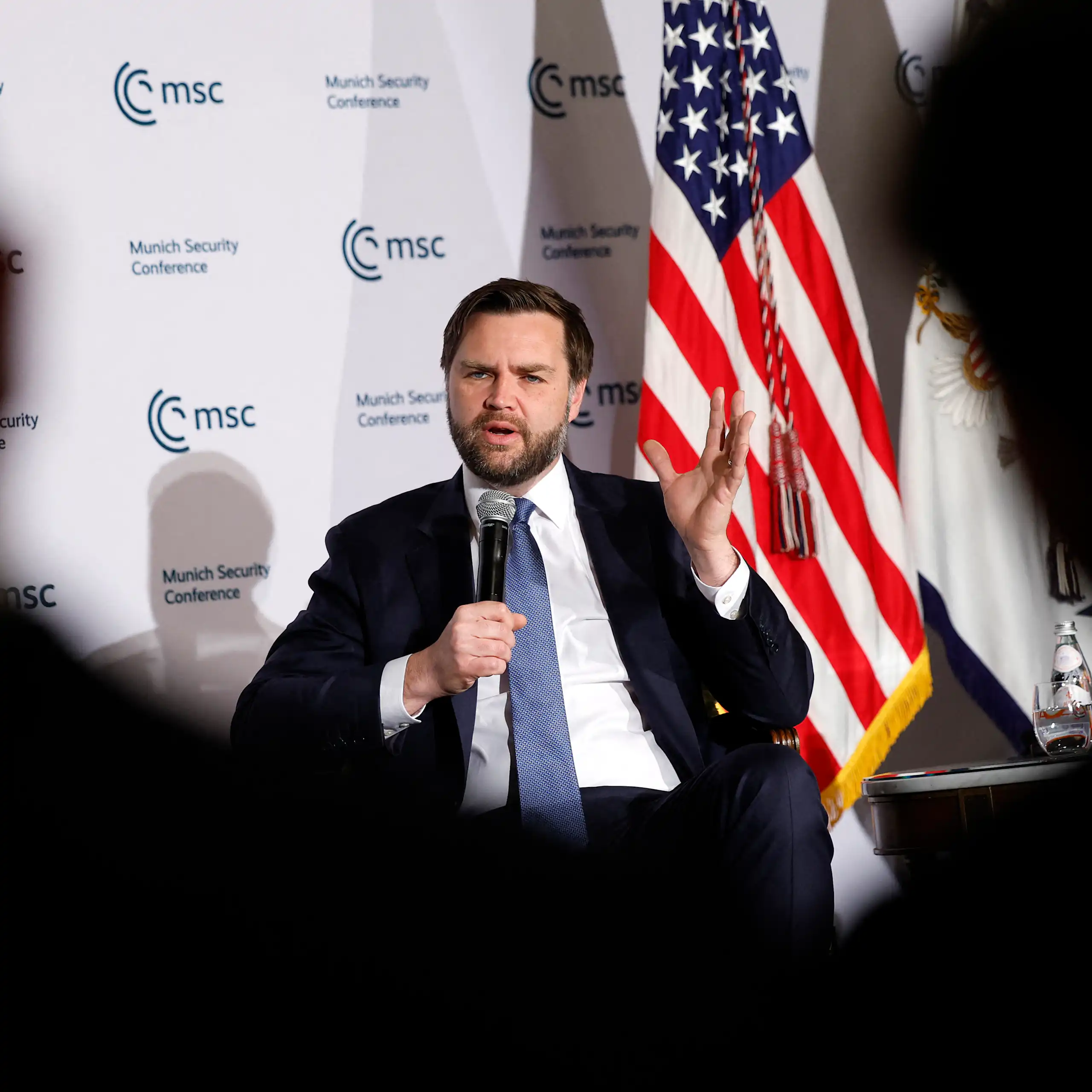 JD Vance assis sur scène tenant un micro devant un drapeau américain