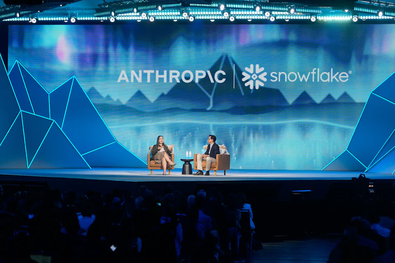 Woman and man sit in brown chairs talking on stage with blue backdrop featuring Anthropic logo.