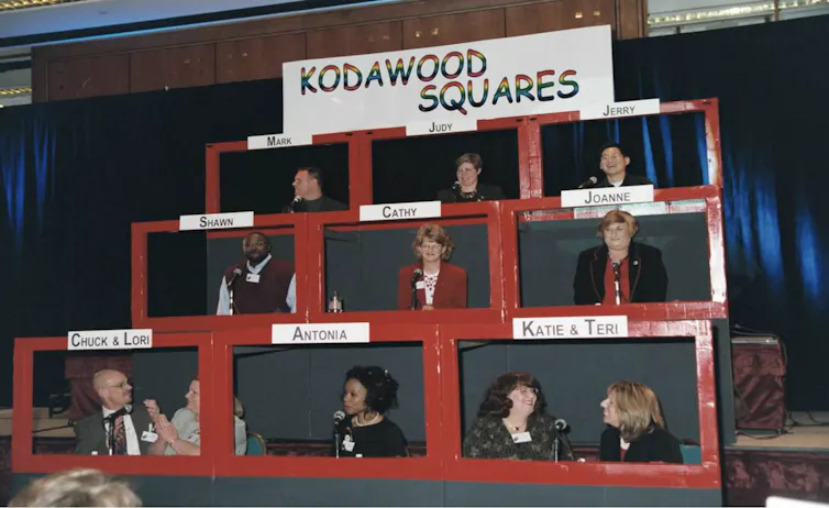 Cómo los empleados LGBTQ de Kodak arriesgaron sus carreras para construir un lugar de trabajo más seguro e inclusivo 2 La gente se sienta detrás de monitores de televisión rojos falsos en el escenario.