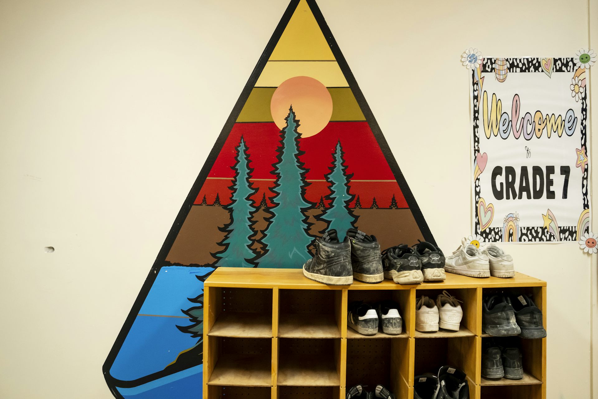 Zapatos para niños en un estante frente a un mural del cielo del amanecer con llamativos árboles de hoja perenne de color naranja, amarillo y rojo, agua y sol.