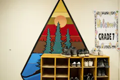 Children's shoes on a shelf in front of a mural of a sunrise sky with bold orange, yellow and red, evergreen trees, water and sun.