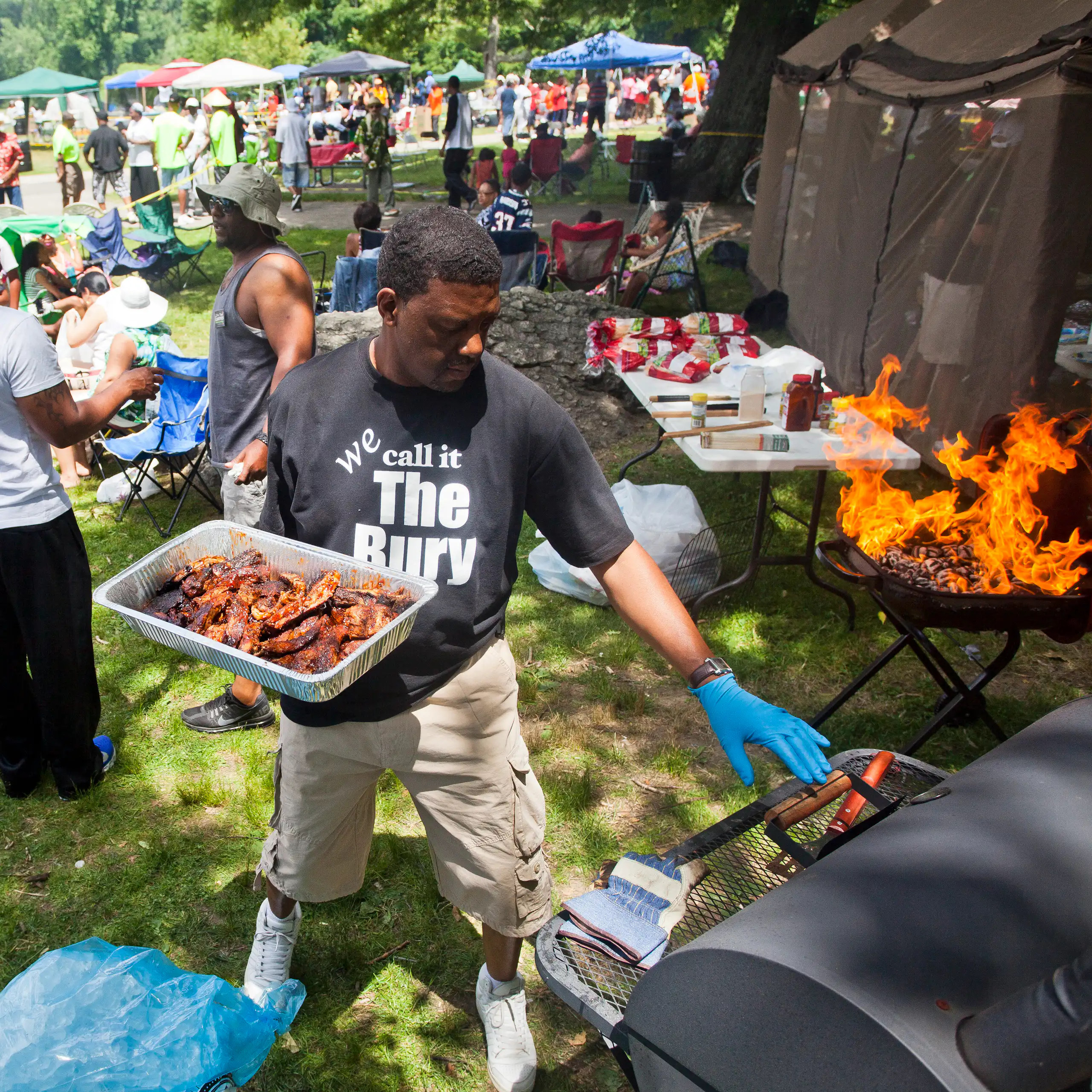 Man holds an aluminum tray of grilled meats in the foreground while a large number of people socialize in the background.