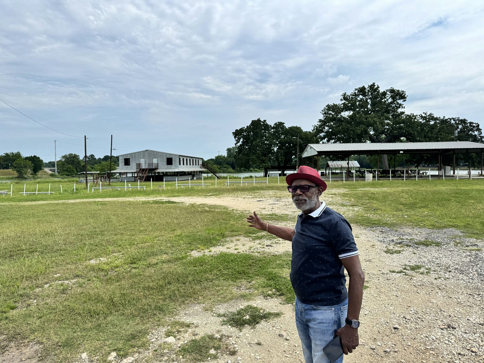 An older Black man wearing a red hat gestures in a field under a cloudy sky.