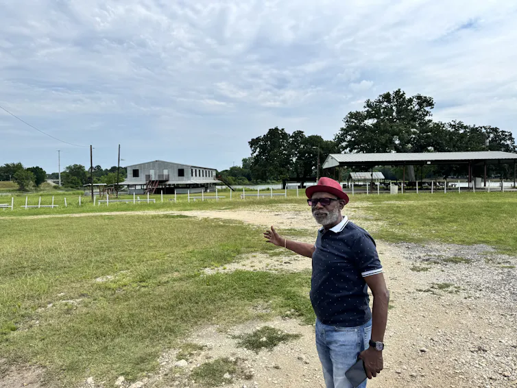 An older Black man wearing a red hat gestures in a field under a cloudy sky.