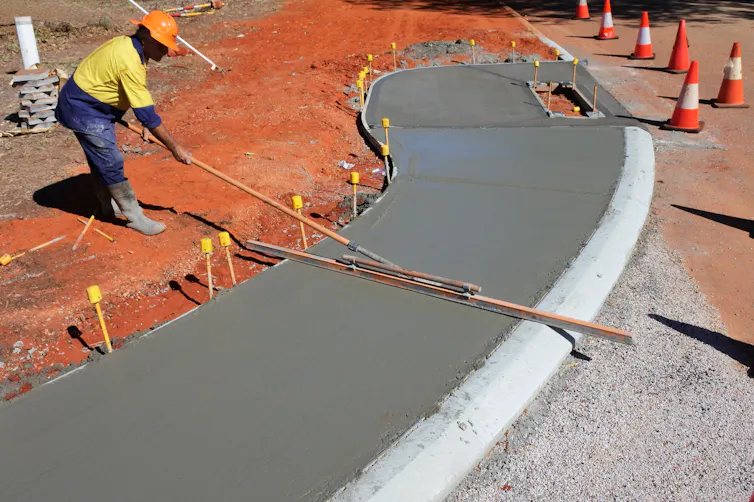 A man spreads concrete on a walkway, wearing high-vis clothing and a hard hat.