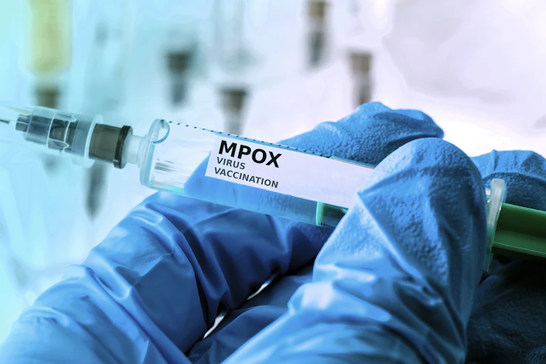 Una mano enguantada sostiene una jeringuillla donde pone "MPOX Vaccine"