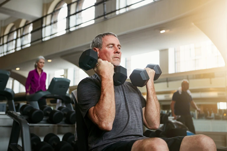An older man sits on a bench in a gym and performs bicep curls with a pair of dumbbells.