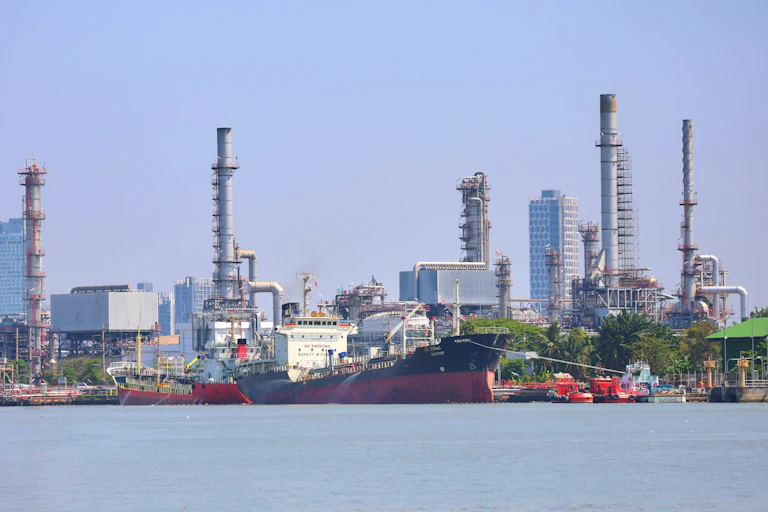 Un barco grande en el mar delante de una zona industrial de procesamiento de petróleo y edificios de fondo.