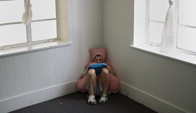 A 10-year-old boy wearing a pink costume sits in the corner of a room and plays on his tablet.