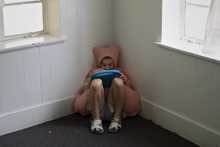 A 10-year-old boy wearing a pink costume sits in the corner of a room and plays on his tablet.
