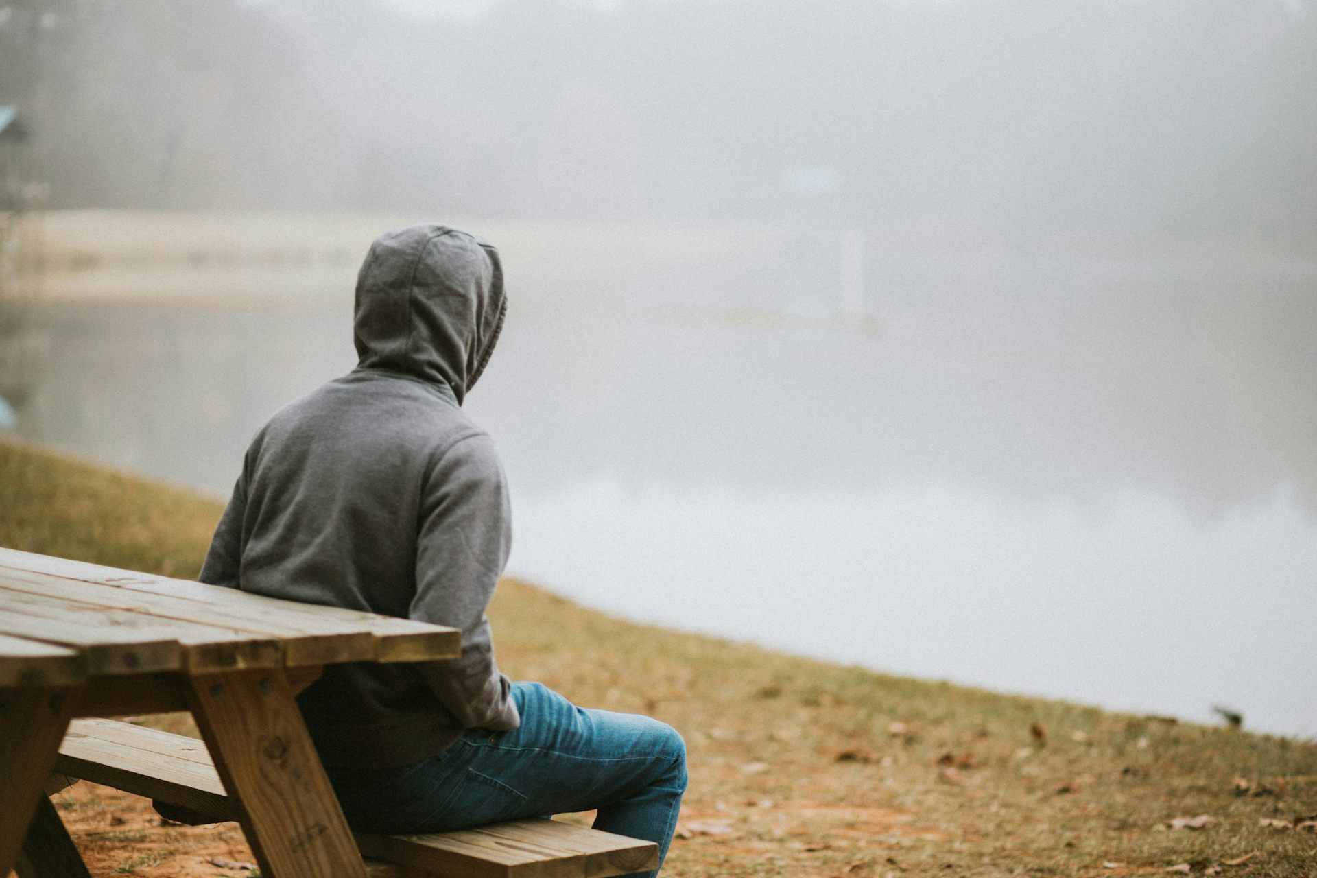 Un jeune homme, vu de dos, vêtu d'un chandail à capuchon, est assis tout seul sur le banc d'une table à pique-nique.