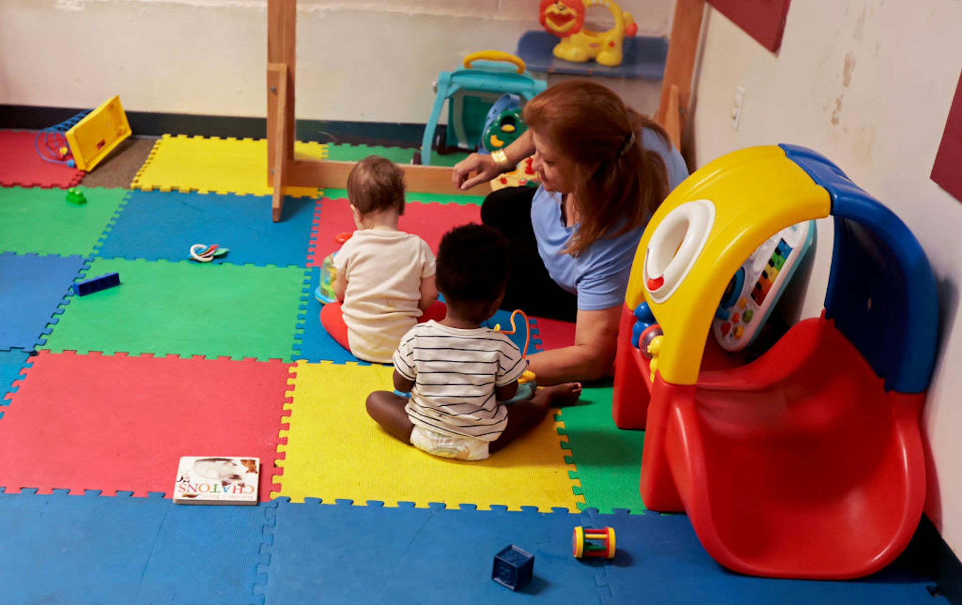 A woman plays with two infants on a colorful mat in a daycare.
