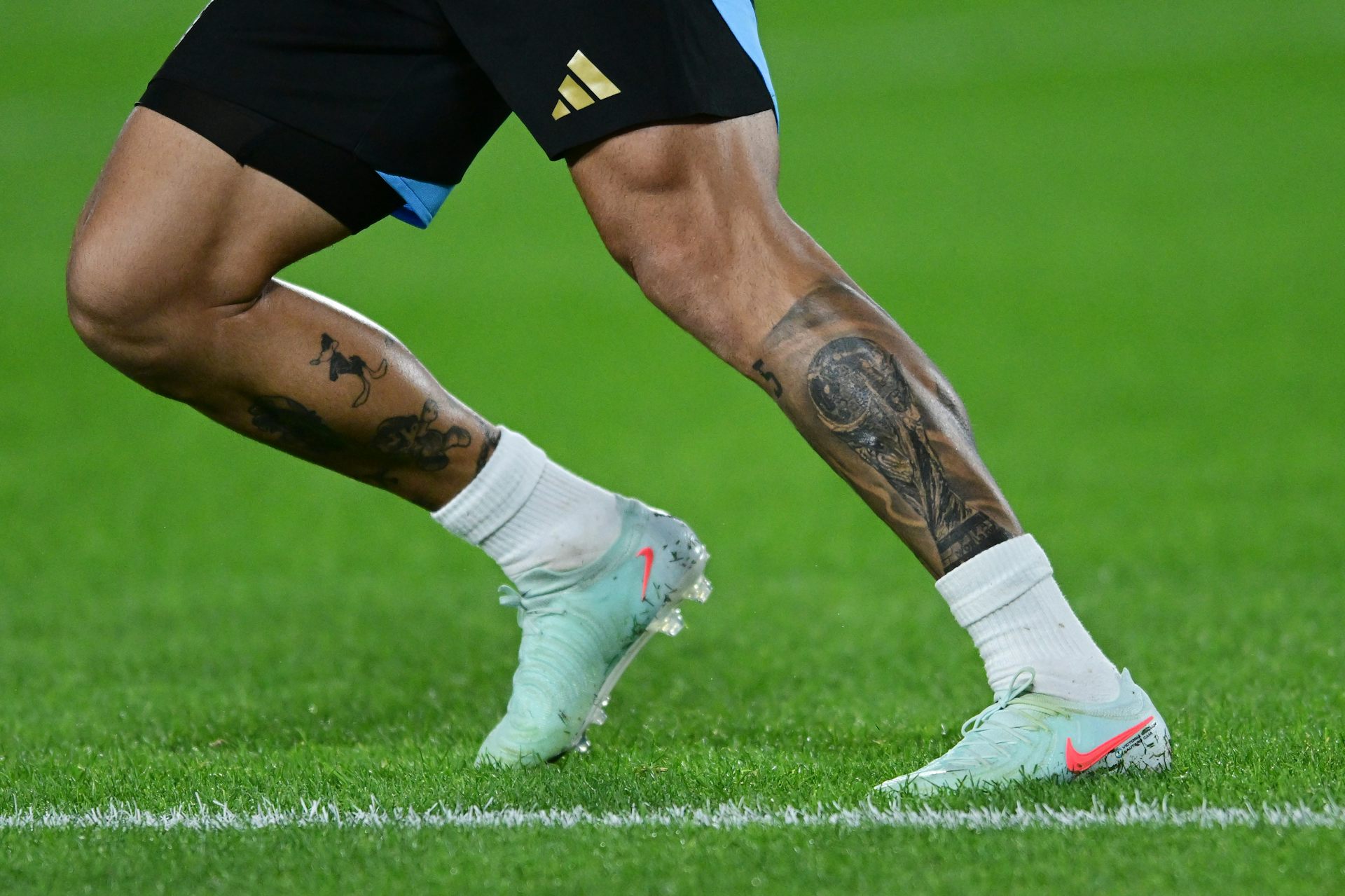 Pernas tatuadas de um jogador de futebol correndo em campo.