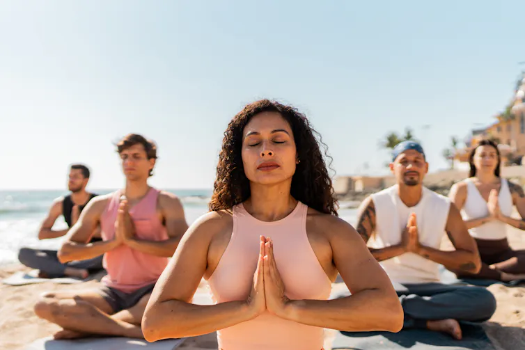 Four people are seated behind a woman leading a meditation practice, all in lotus pose with their hands folded in namaste.