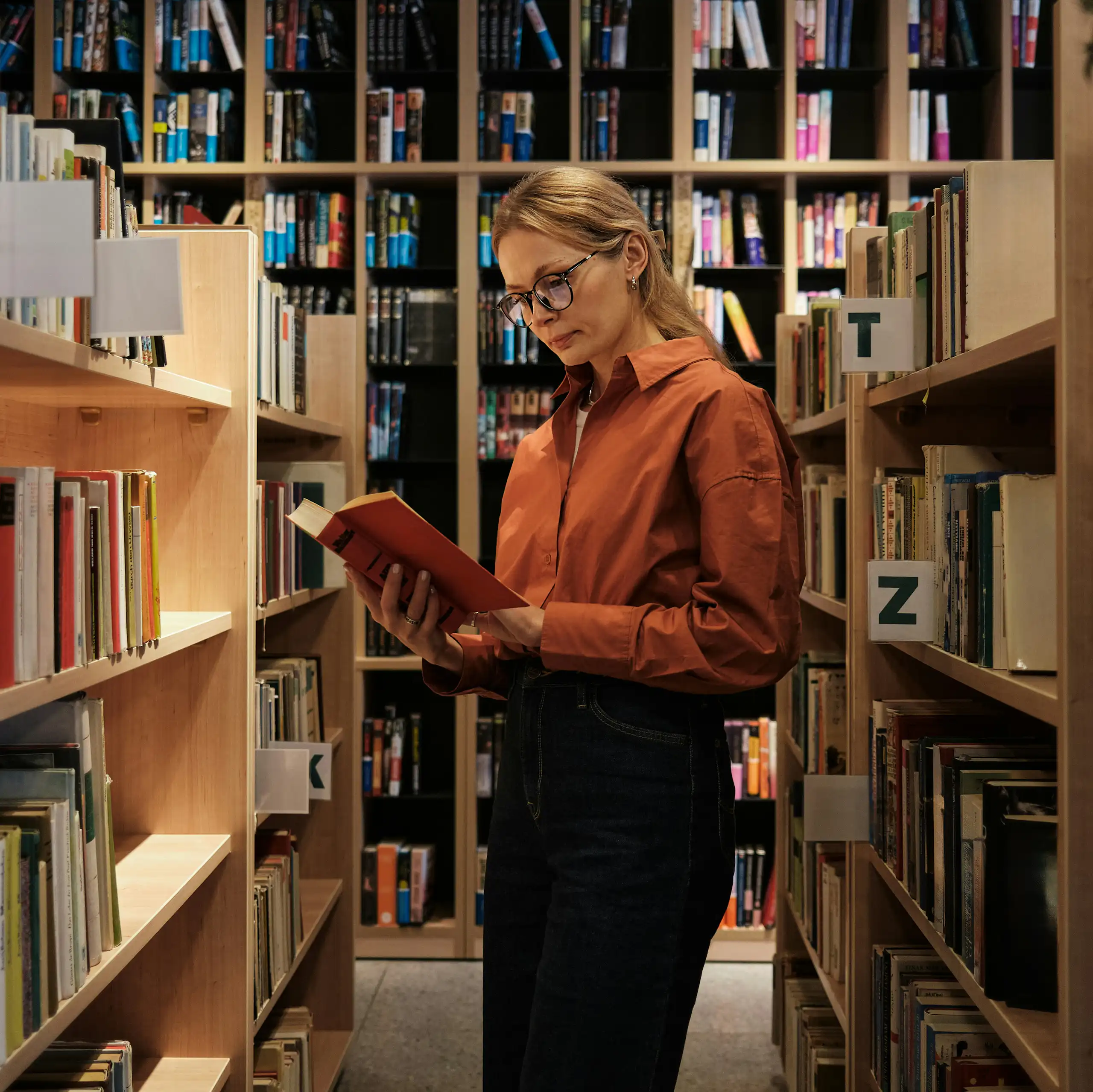 A young woman stands between shelves in a library reading a book