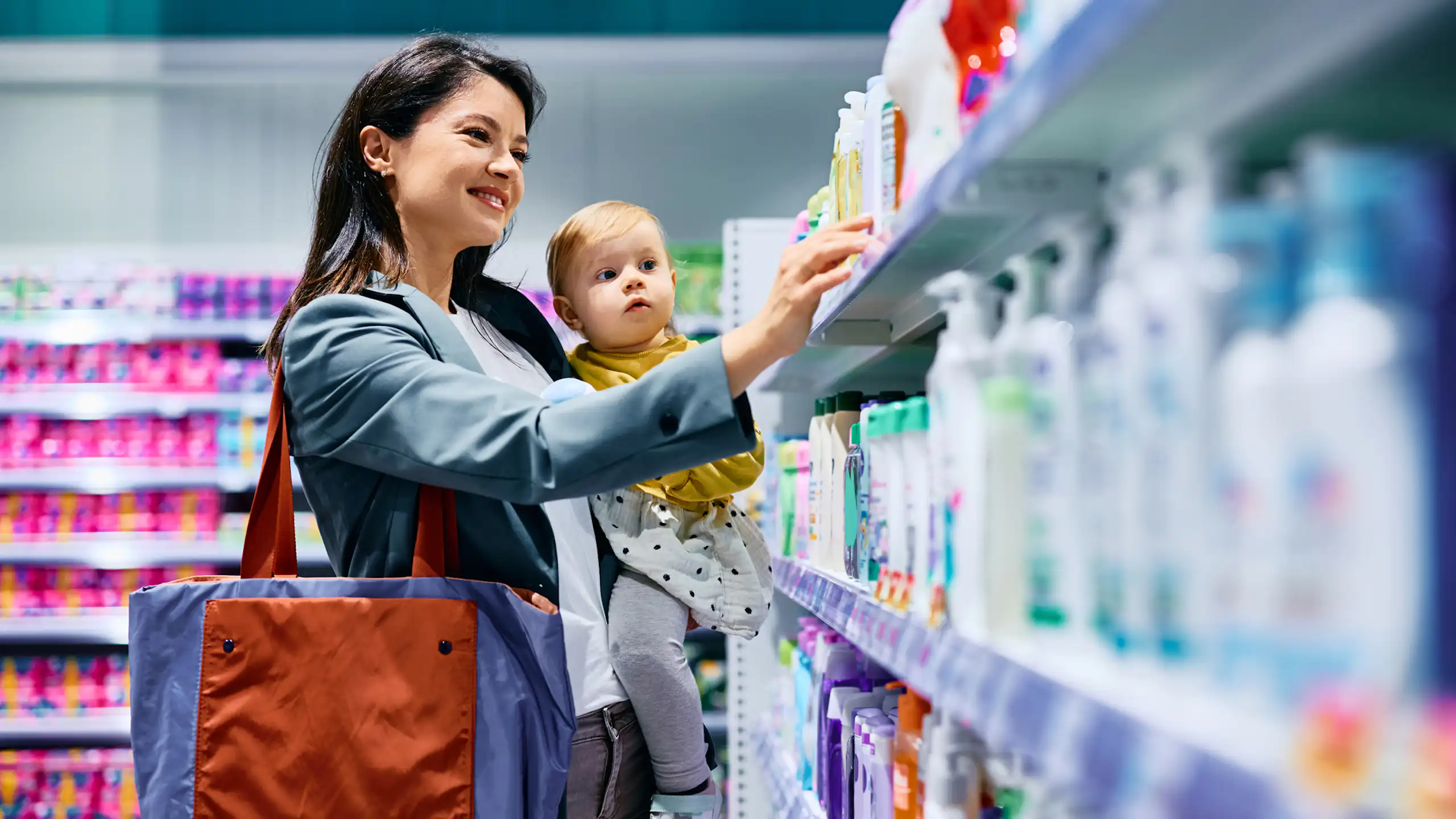 Mother with young child in arm choosing products on pharmacy shelf.
