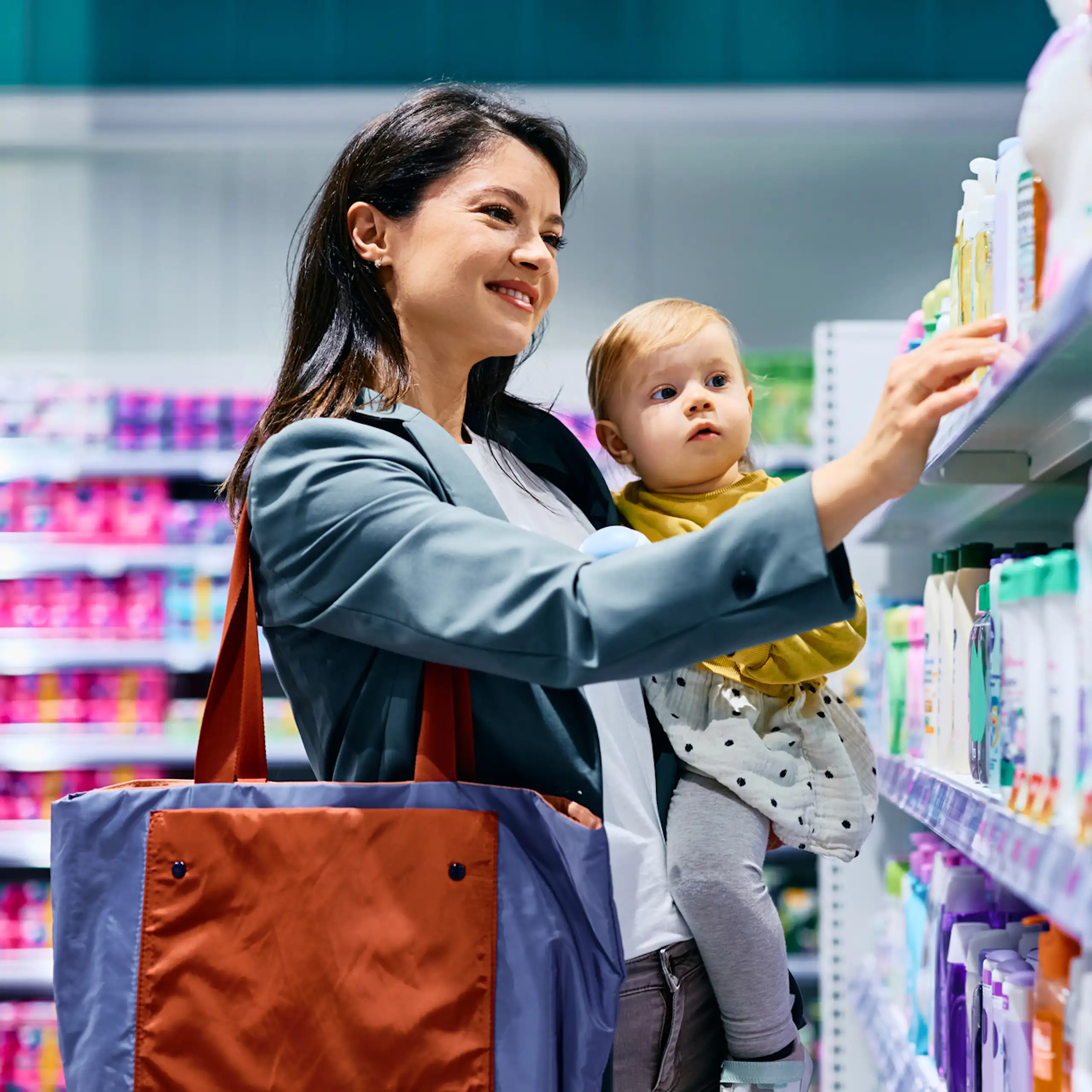 Mother with young child in arm choosing products on pharmacy shelf.