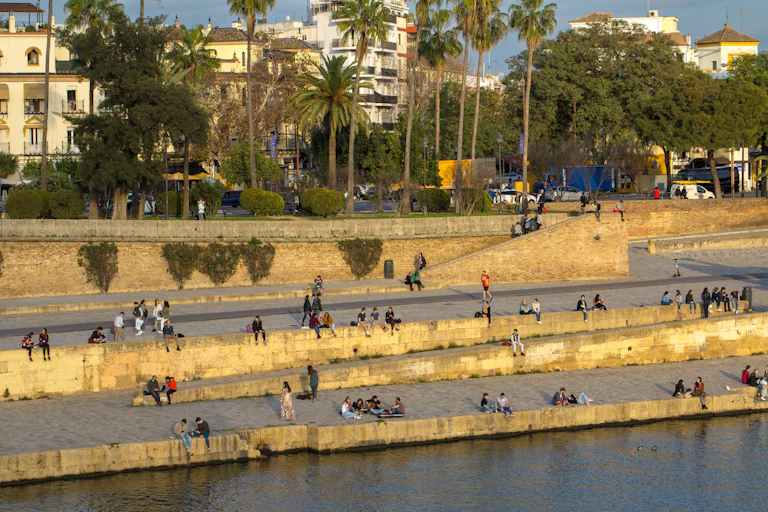 Una escena animada a lo largo de las orillas del río iluminadas por el sol en Sevilla, donde la gente se relaja en múltiples terrazas de piedra, rodeada de arquitectura histórica y palmeras mecedoras