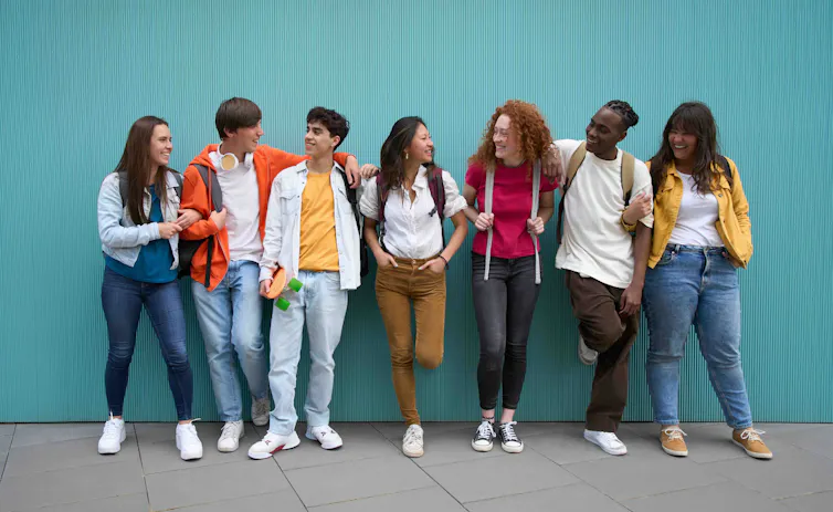 A group of young friends are leaning against a blue wall and smiling at each other.