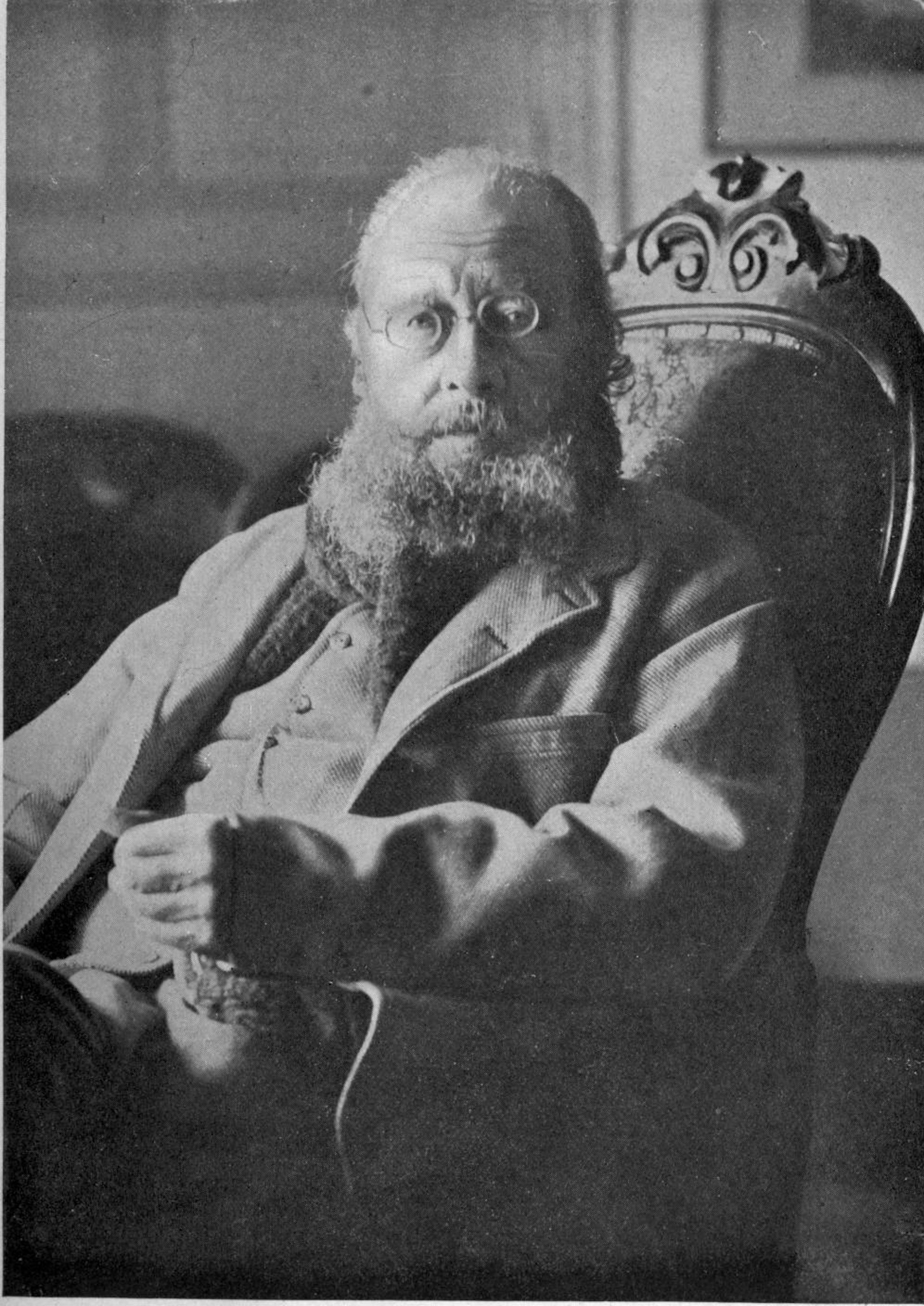 An old man with spectacles and a big bush beard sitting in an ornate chair from the 19th century.