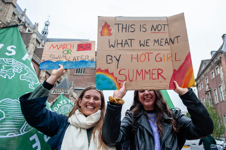 Two women hold signs, one reading 'This is not what we meant by Hot Girl Summer', and 'March now or swim later'