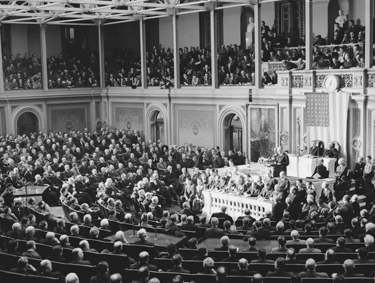 A large chamber filled with people seated in curved rows, being addressed by a an in the front of the room.
