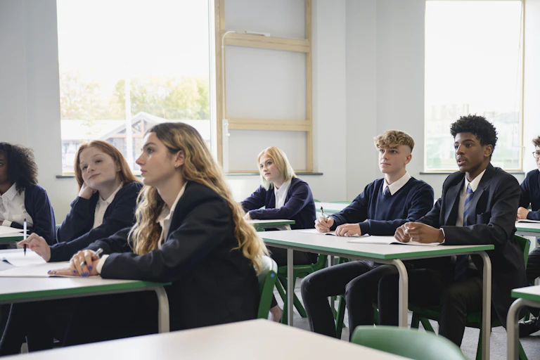A group of high students wearing uniforms, sitting side by side at desks, and listening to an off-camera teacher.