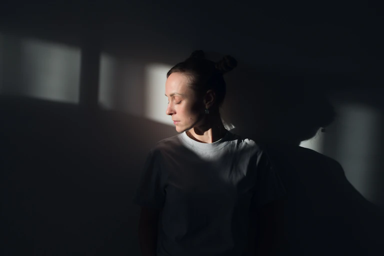A woman stands against a white wall, in the rays of sunlight, with her eyes closed.