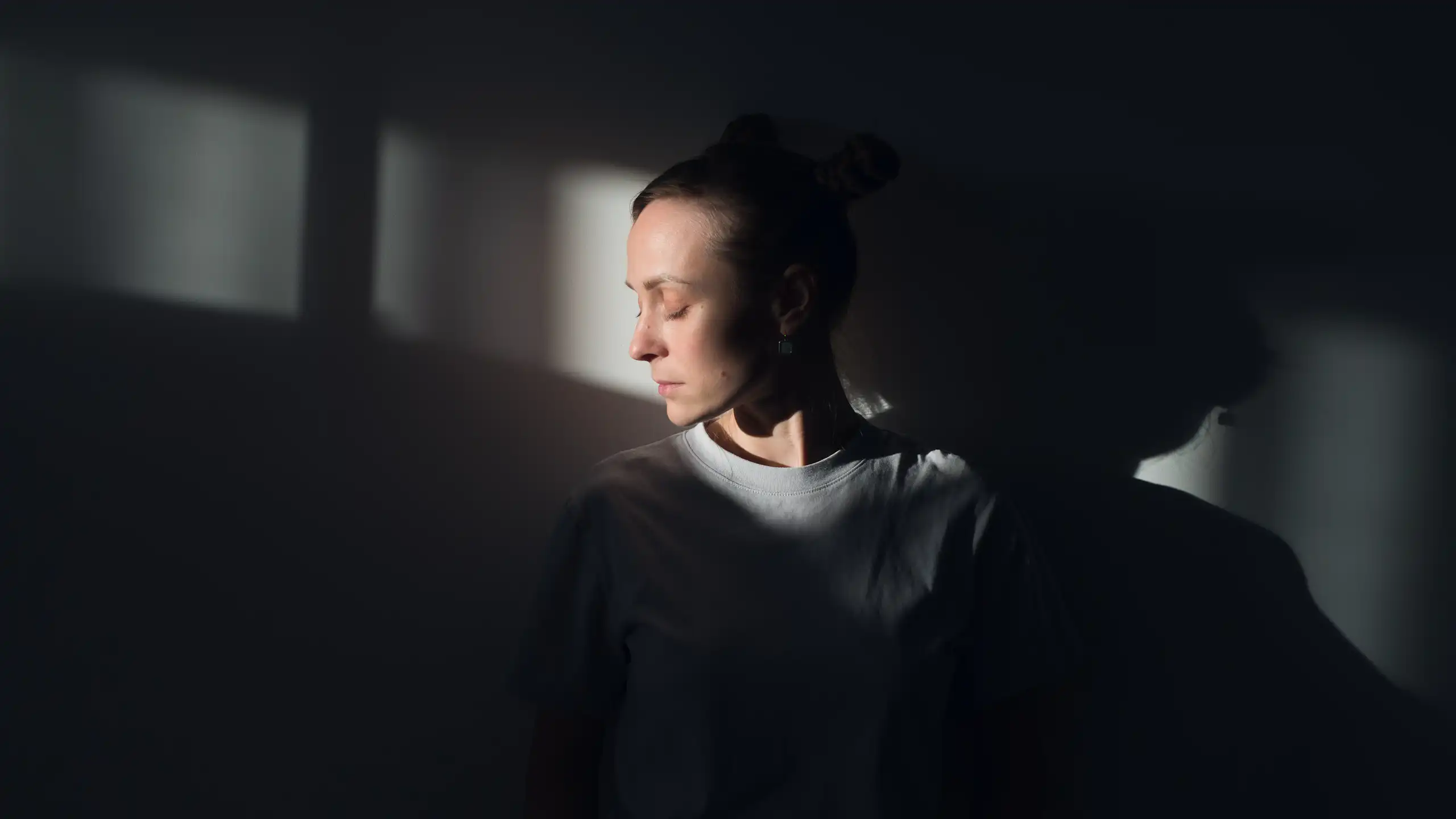 A woman stands against a white wall, in the rays of sunlight, with her eyes closed.