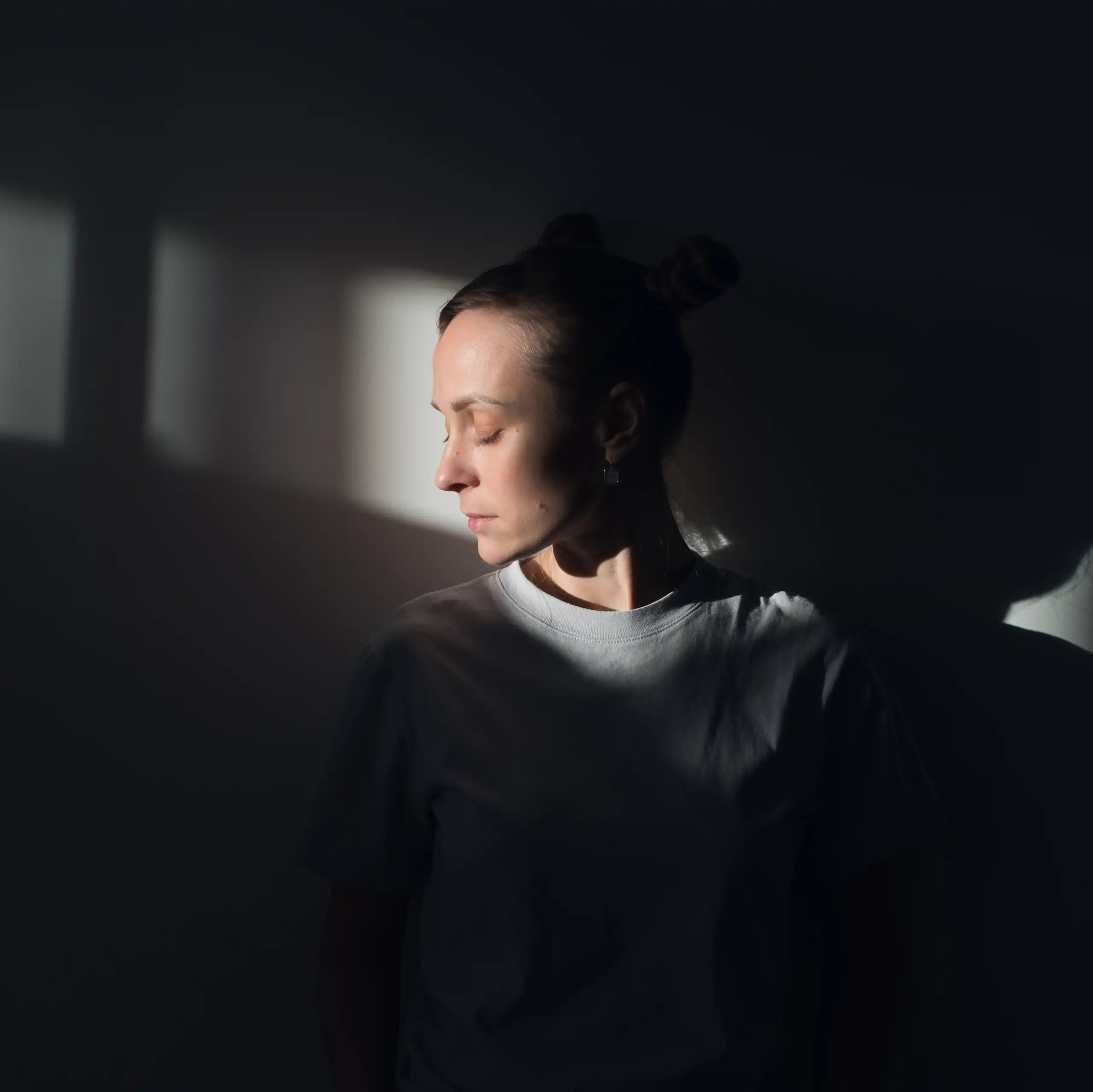 A woman stands against a white wall, in the rays of sunlight, with her eyes closed.