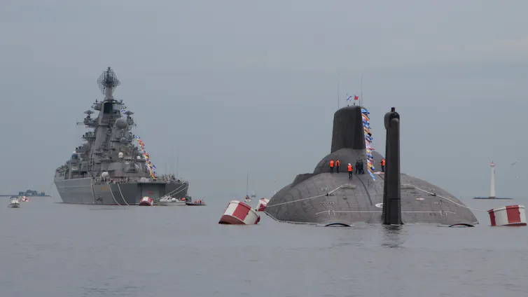 A Russian nuclear submarine surfaces alongside a Russian Navy warship.