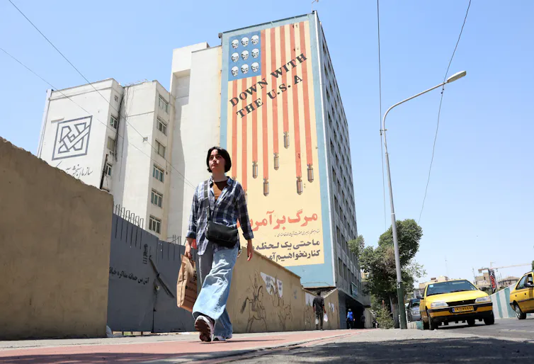 A young woman walks past an anti-US mural, June 2026