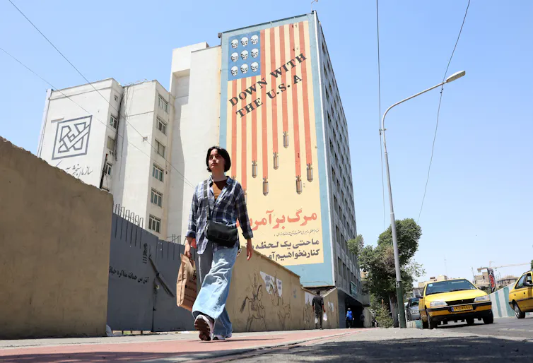 A young woman walks past an anti-US mural, June 2026