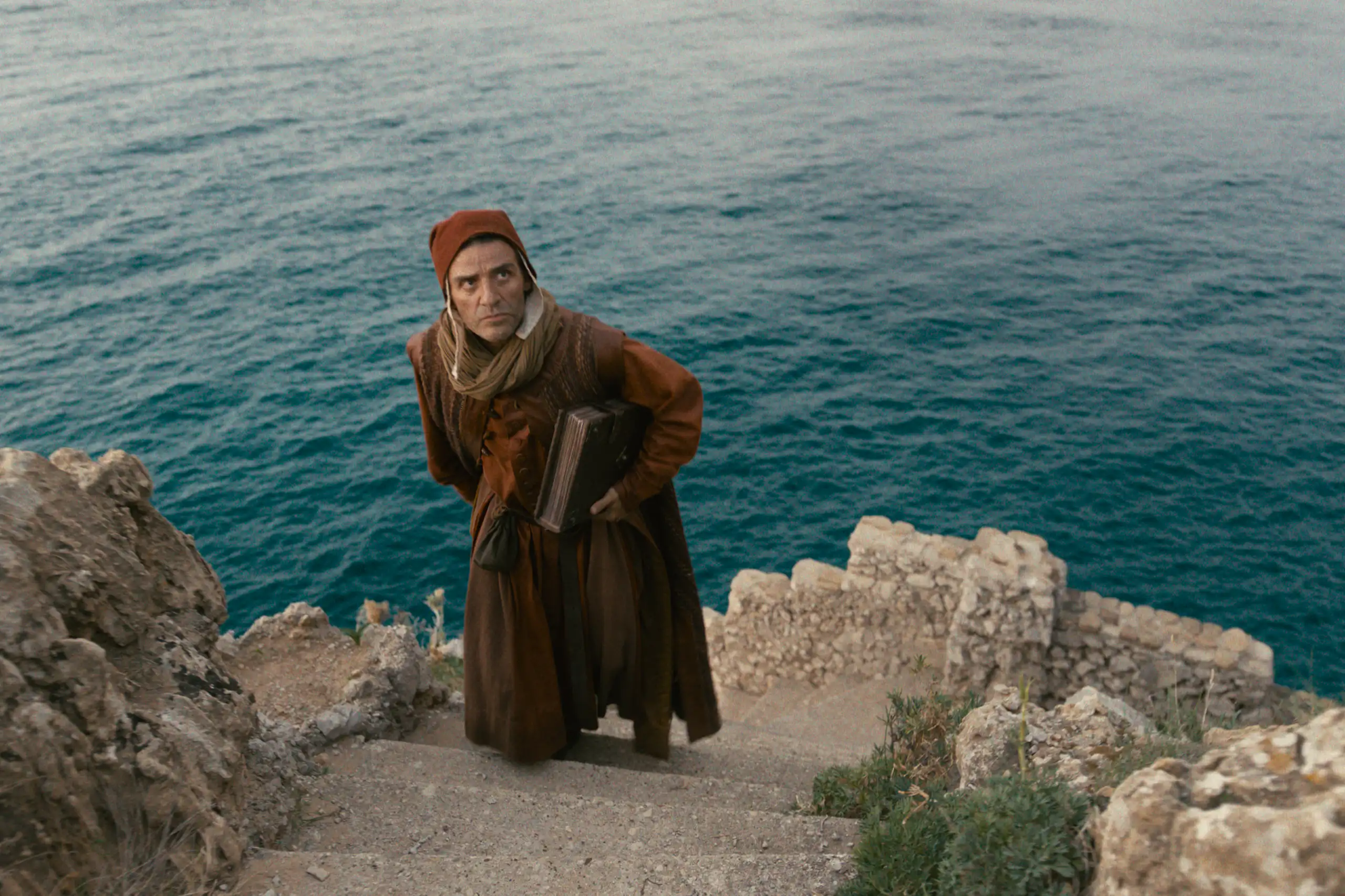 A man in medieval dress climbing some stone steps with the sea in the background.