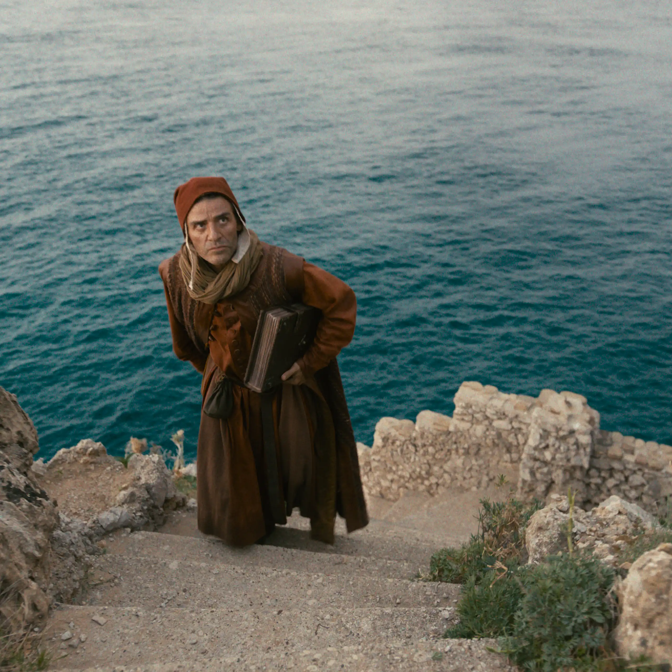 A man in medieval dress climbing some stone steps with the sea in the background.