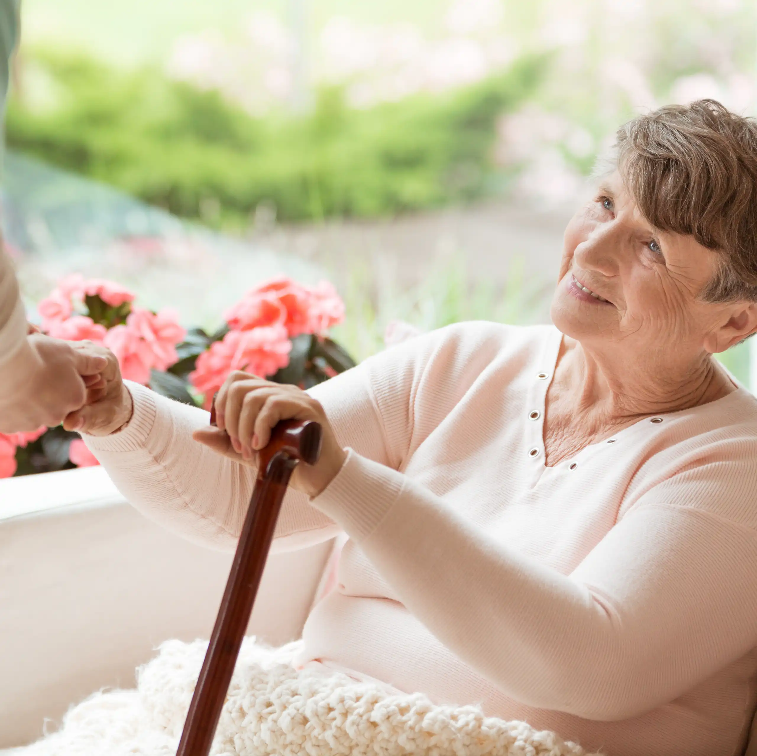 Une femme âgée assise dans un fauteuil tient une canne à la main et discute avec une soignante.