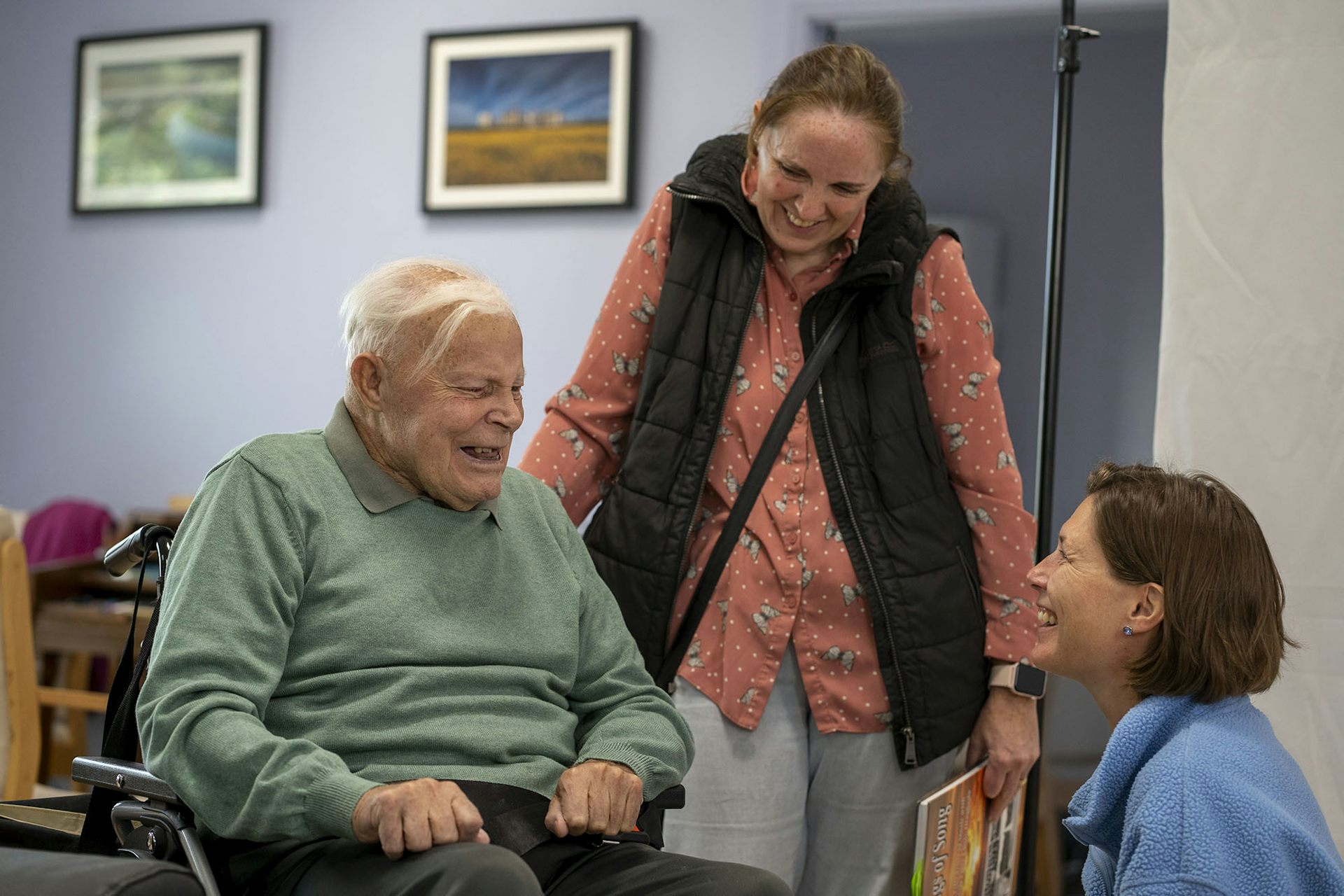 Woman sitting in front of older, wheelchair using, man. Another woman stands with her hand on the back of the man's wheelchair. The three people are laughing together.