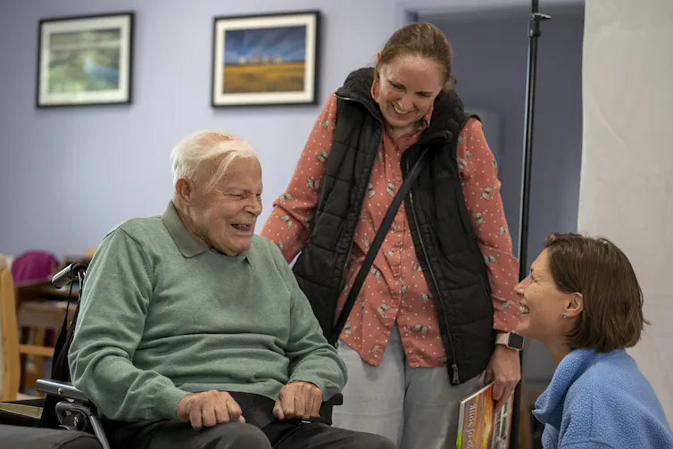 Woman sitting in front of older, wheelchair using, man. Another woman stands with her hand on the back of the man's wheelchair. The three people are laughing together.