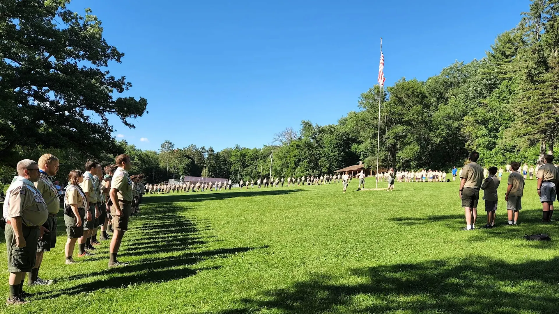 People of all ages in beige uniforms stand in a line in a green field on a sunny day before an American flag on a pole.