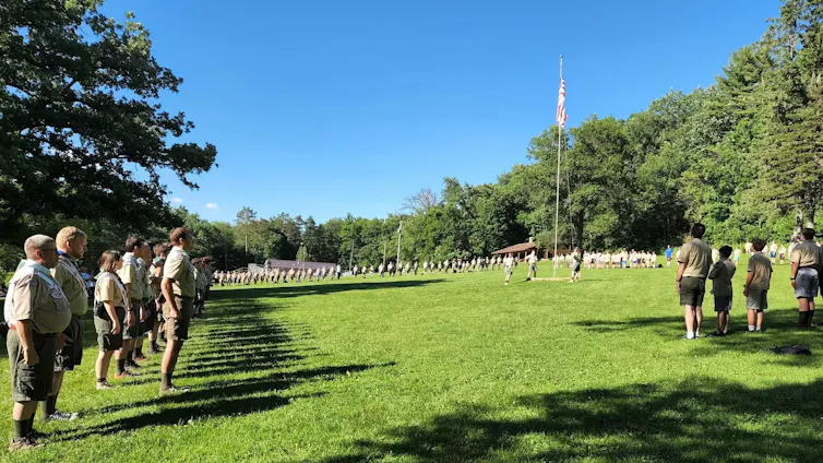 People of all ages in beige uniforms stand in a line in a green field on a sunny day before an American flag on a pole.