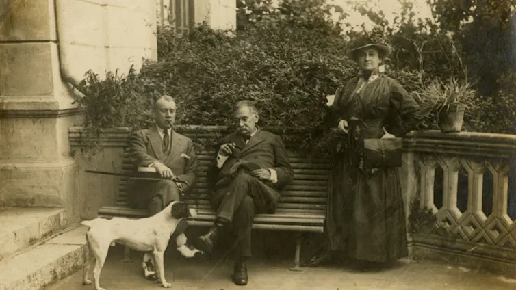 Fotografía de dos hombres sentados y una mujer de pie de principios del siglo XX. Un perro blanco les mira.